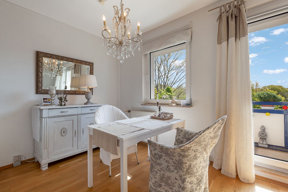 Helles Zimmer mit Esstisch, zwei Stühlen, Kronleuchter und Zugang zum Balkon.