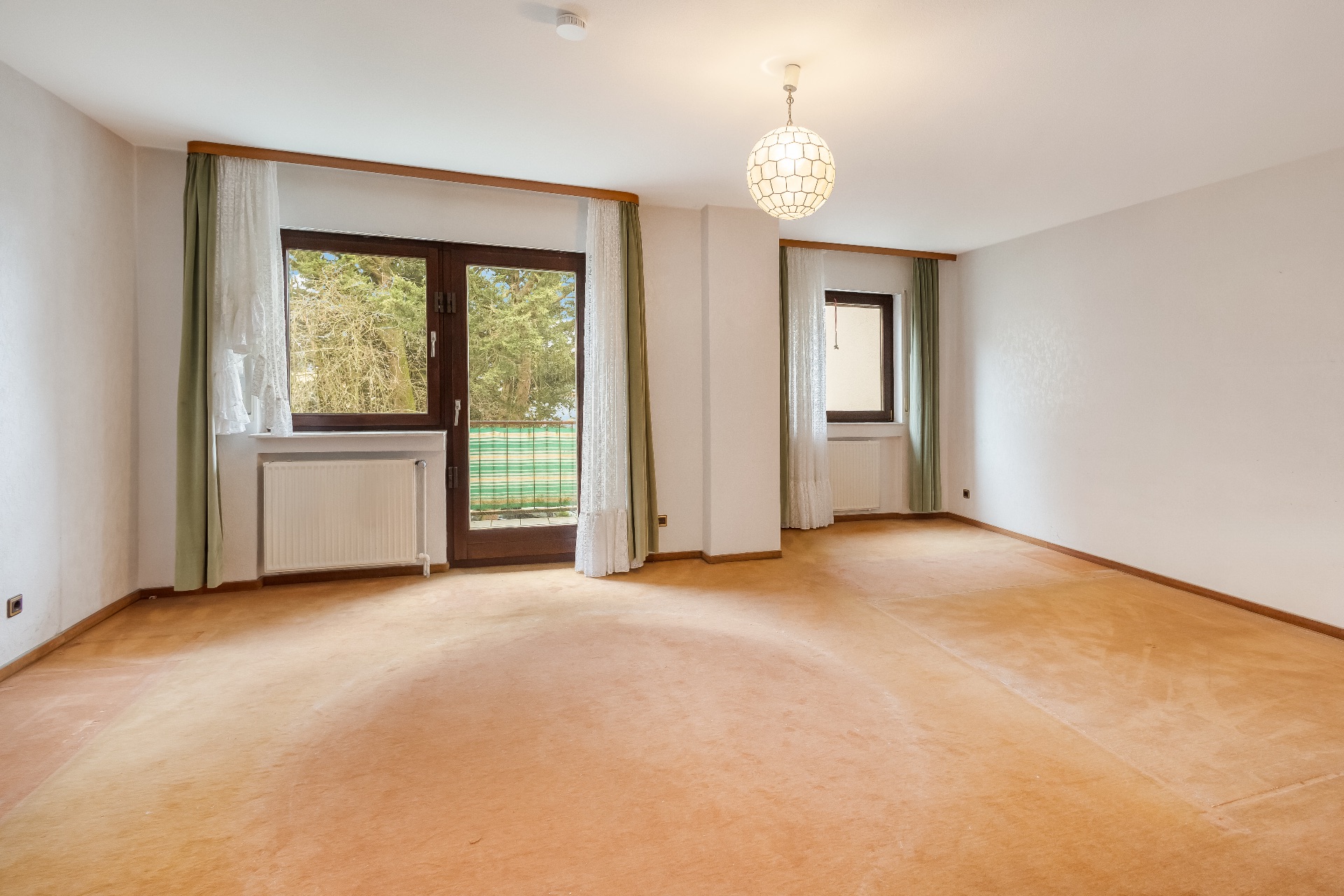 Leeres Zimmer mit Teppichboden, zwei Fenstern, Balkontür und weißer Hängelampe.