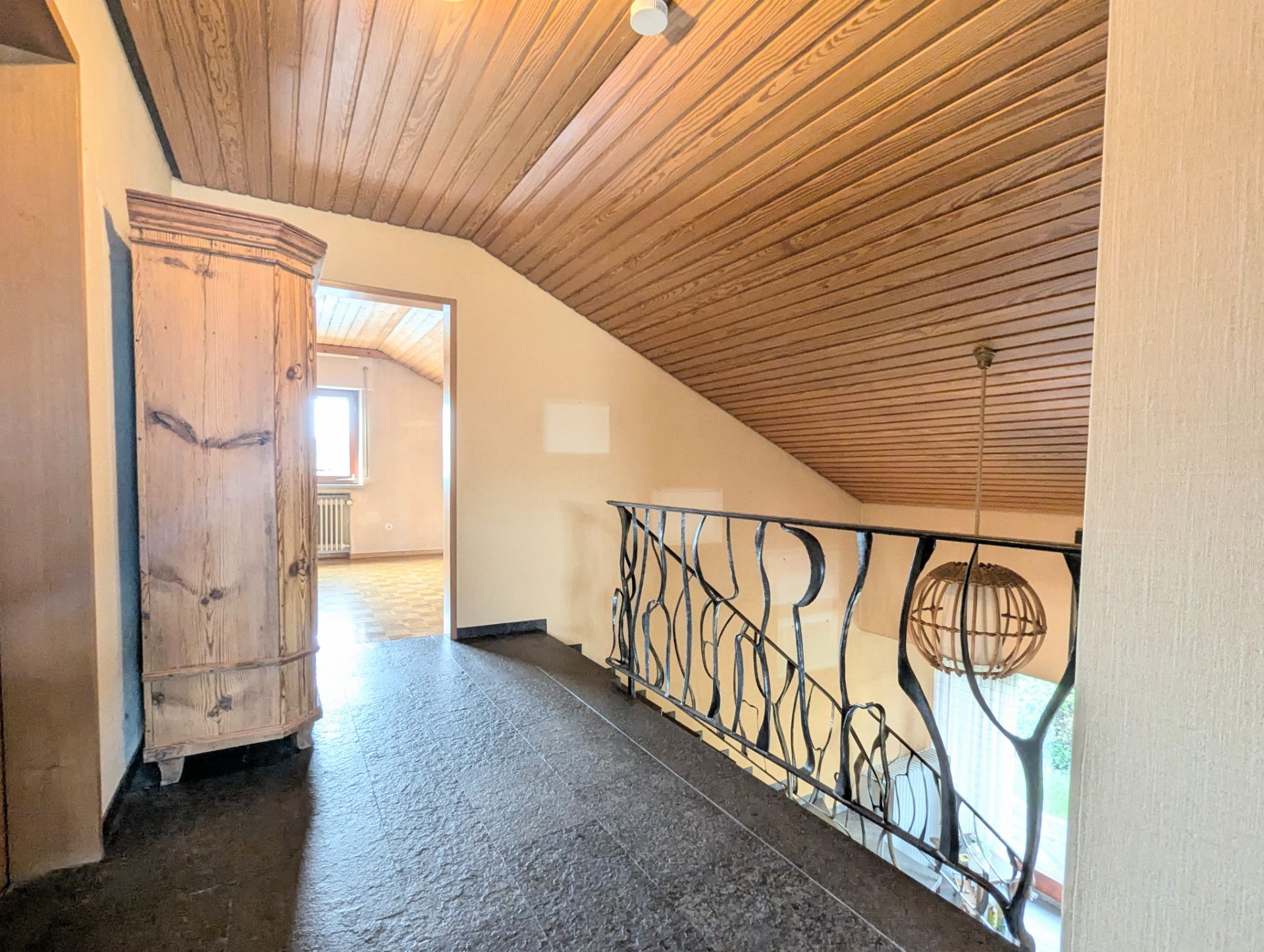 Dachgeschossflur mit Holzdecke, Schrank, Geländer und Blick in ein Zimmer mit Fenster.