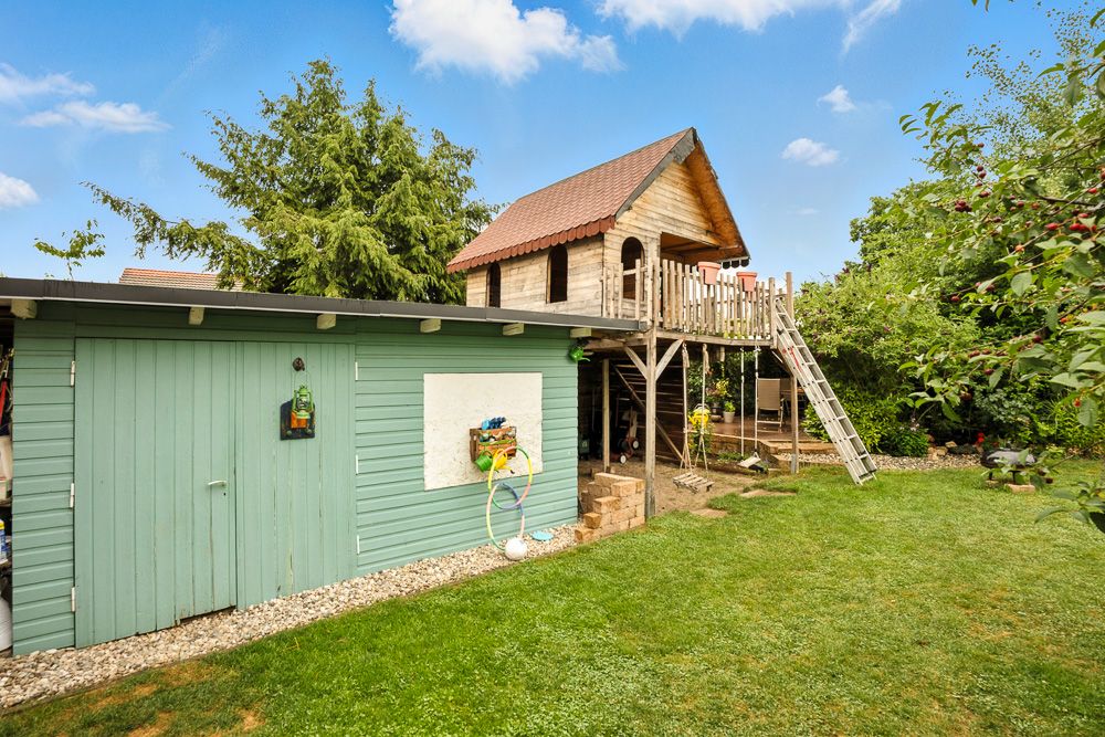 Garten mit grünem Schuppen und großem Holzspielhaus auf Stelzen mit Rutsche.