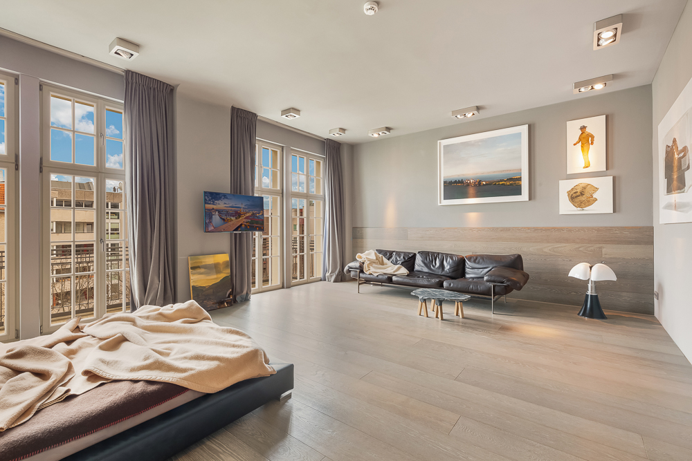 Helles Wohnzimmer mit großen Fenstern, grauen Vorhängen, Sofa, Bett und Wandkunst.