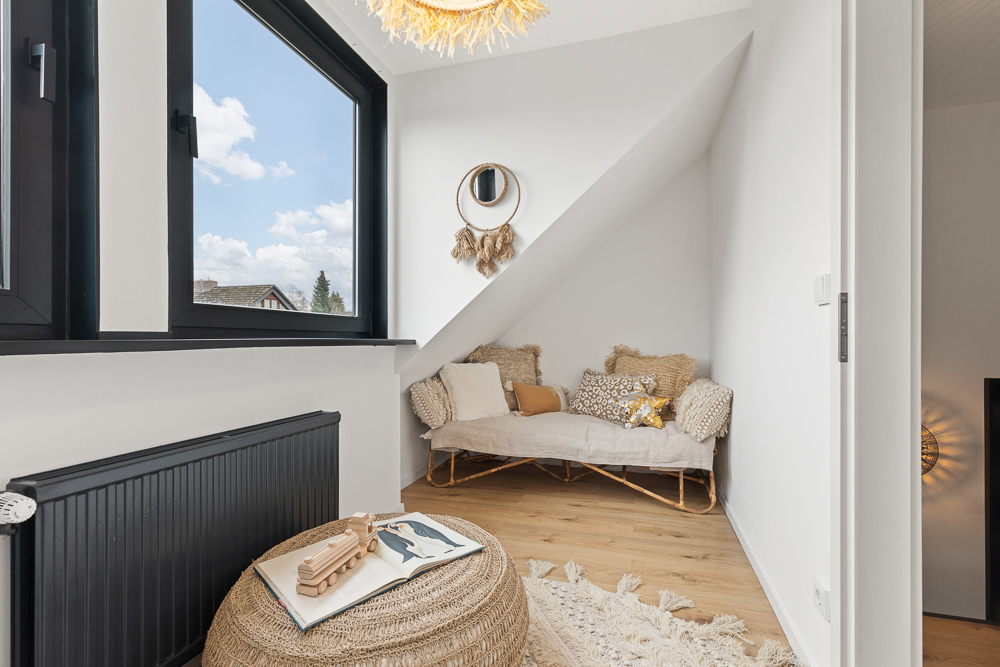 Gemütliche Ecke mit Sofa, Kissen und Pouf vor großem Fenster unter einer Dachschräge.