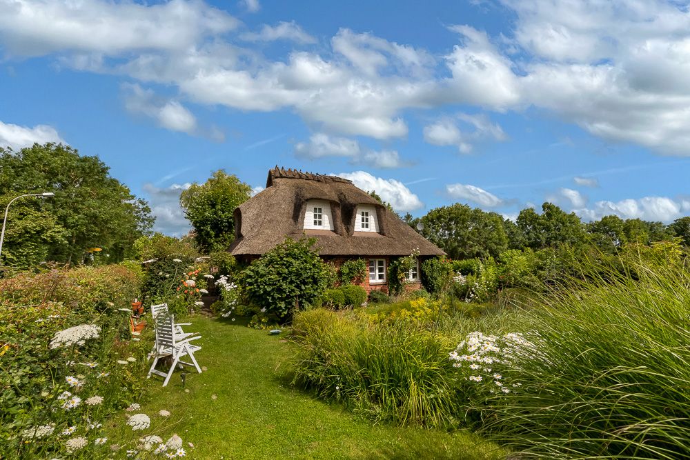 Kleines Haus mit Reetdach im blühenden Garten, zwei weiße Gartenstühle auf der Wiese.