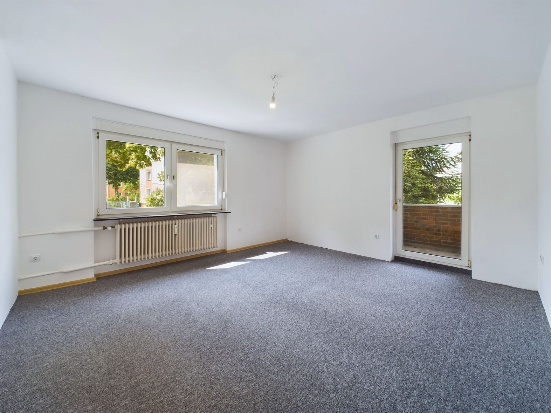 Leeres Zimmer mit Teppichboden, großem Fenster, Heizung und Tür zum Balkon.