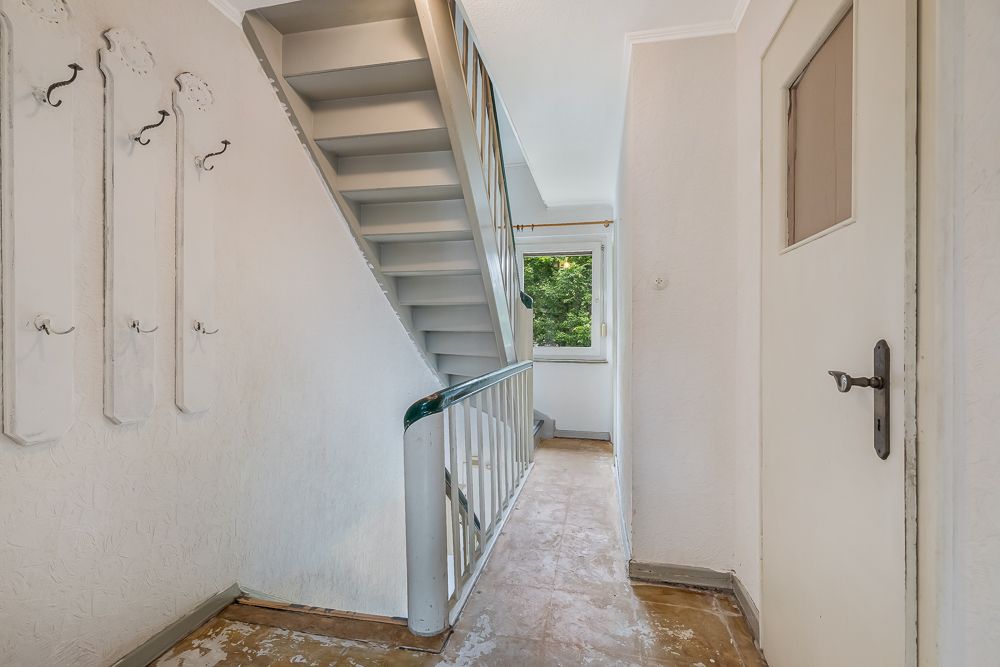 Flur mit Treppe, Fenster, mehreren Wandhaken und einer weißen Tür.
