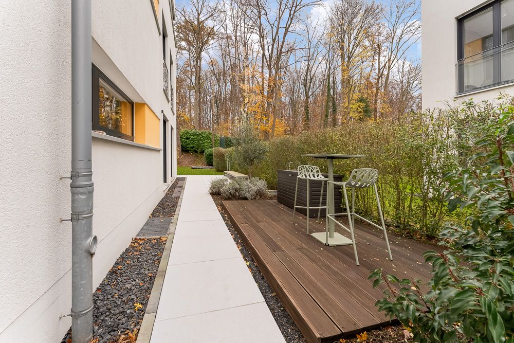 Moderner Außenbereich mit Holzterrasse, zwei Stühlen und Tisch, umgeben von Hecken und Bäumen.