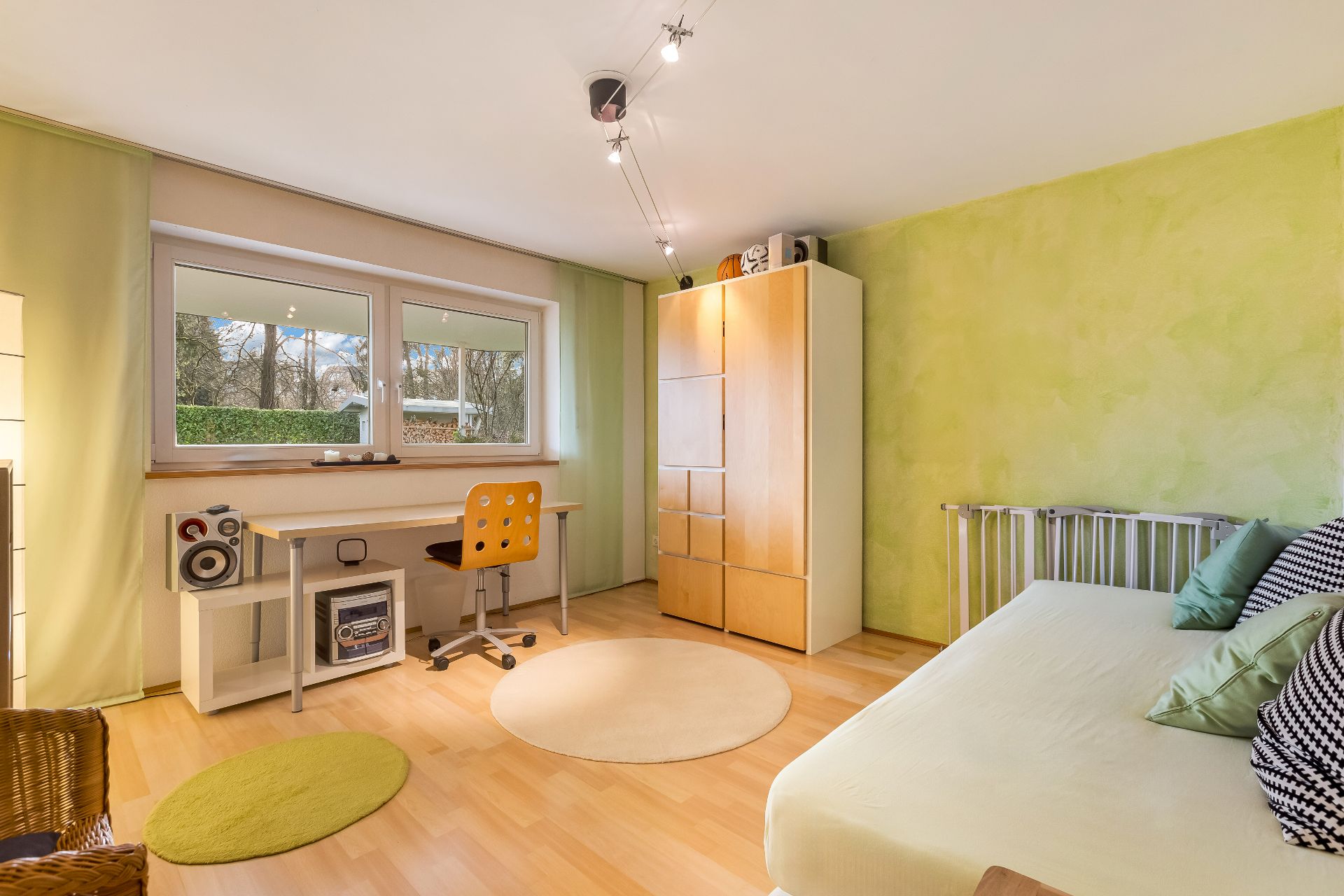 Helles Zimmer mit Bett, Schreibtisch, Stuhl, Schrank und großen Fenstern.