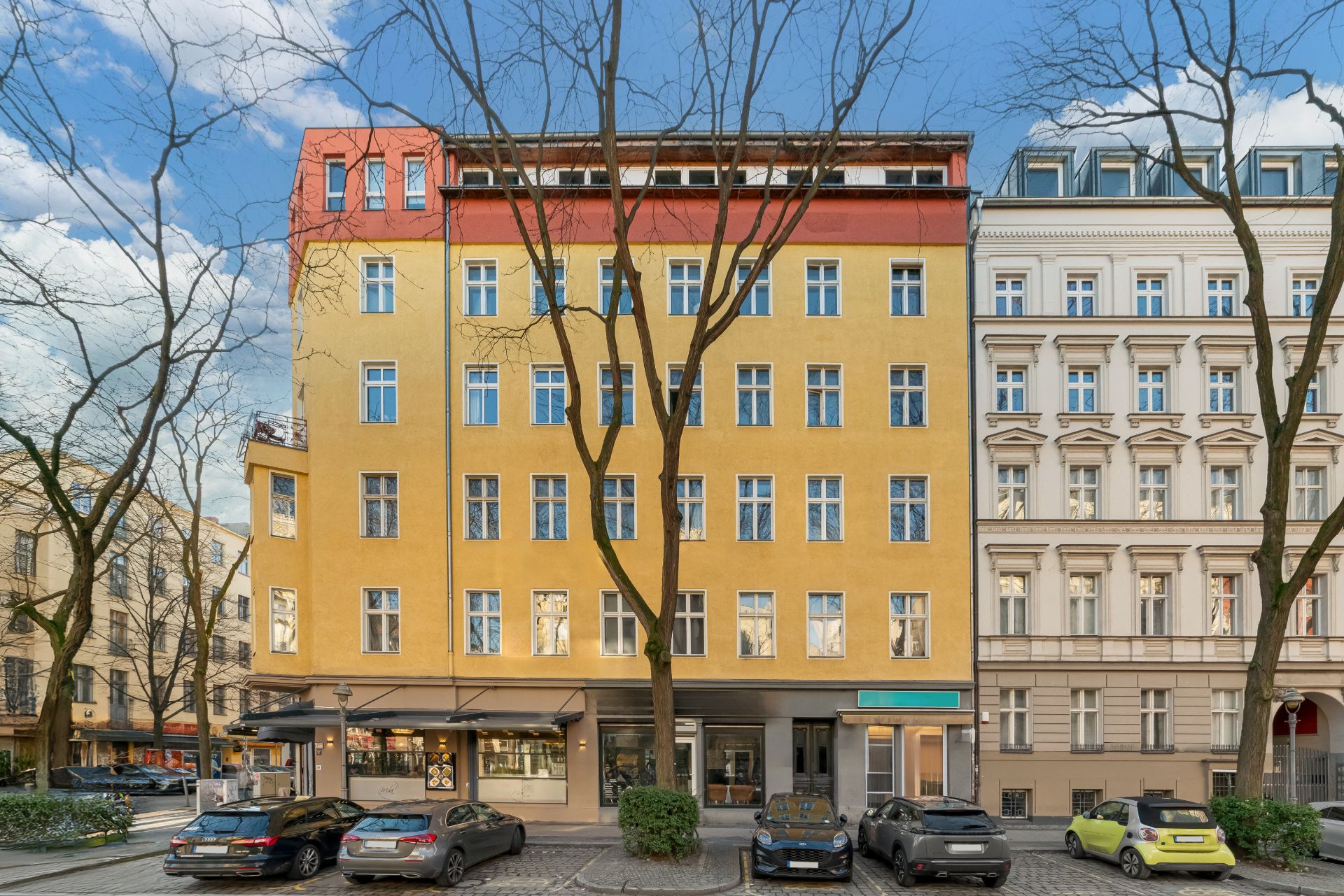 Gelbes Wohnhaus mit vielen Fenstern und Bäumen davor an einer Straße mit geparkten Autos.