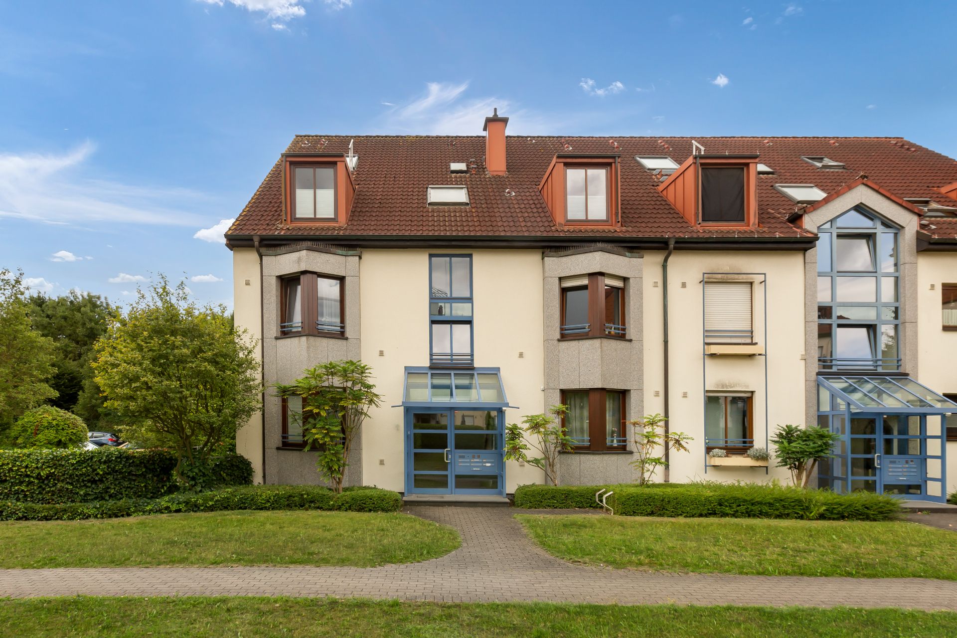 Dreistöckiges Wohnhaus mit roten Dachziegeln und Glas-Eingangstür, umgeben von Rasen und Bäumen.
