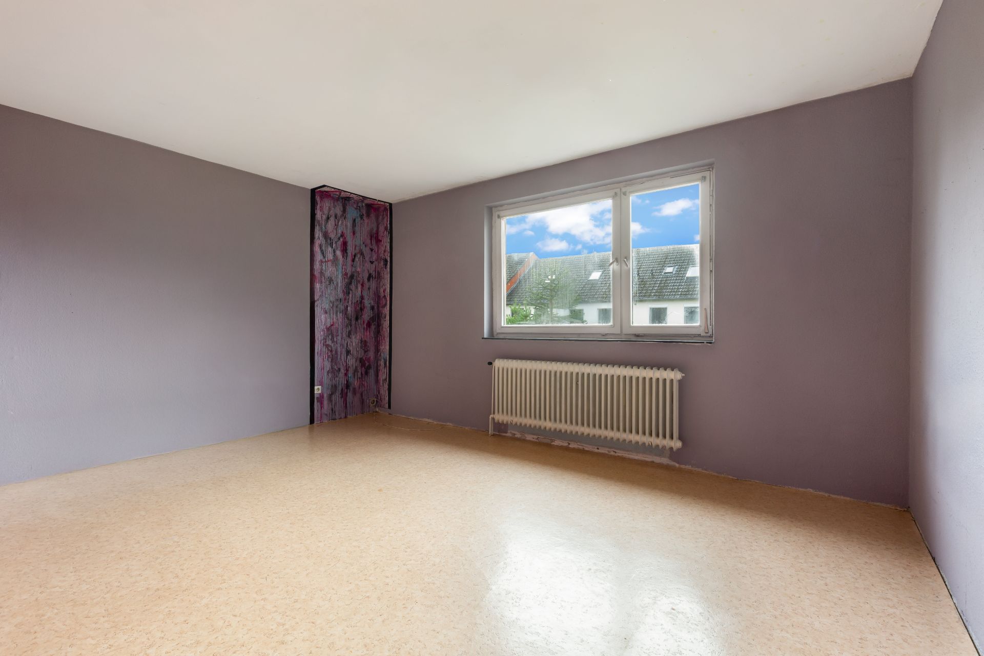 Leeres Zimmer mit lila Wänden, großem Fenster und Heizkörper darunter.
