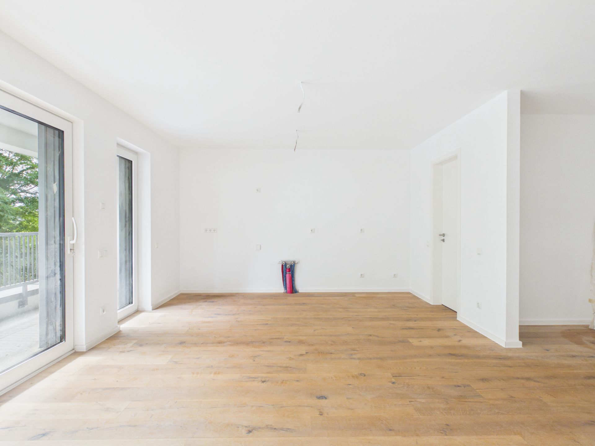 Leerer, heller Raum mit Holzboden und großen Fenstern zur Terrasse.