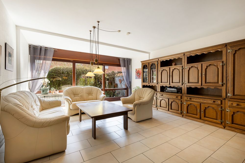 Wohnzimmer mit hellen Ledersofas, Holztisch, großer Schrankwand und bodentiefem Fenster.