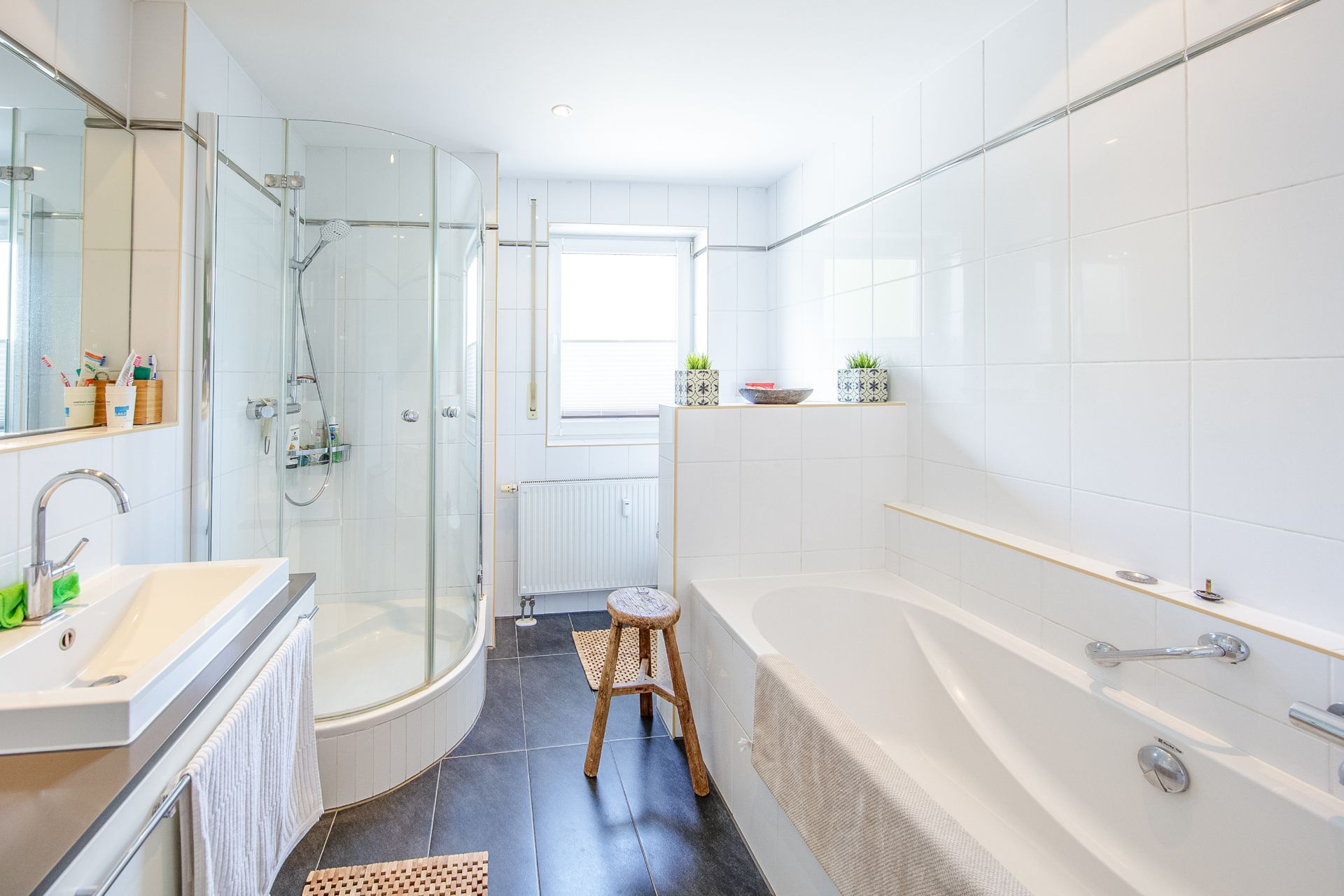 Modernes Badezimmer mit Dusche, Badewanne, Waschbecken und Fenster. Ein Hocker steht neben der Badewanne.