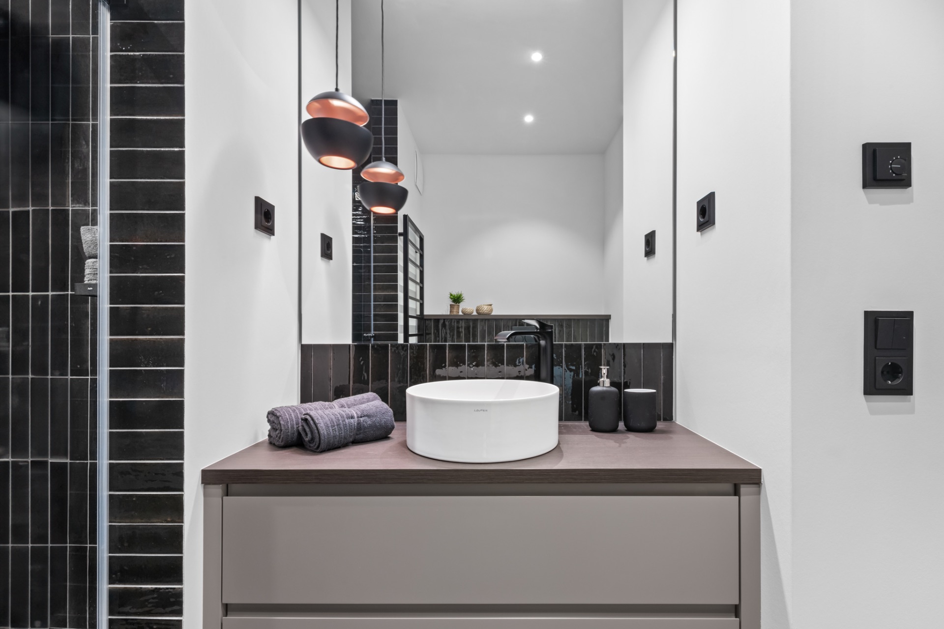 Modernes Badezimmer mit rundem Waschbecken, Spiegel und schwarzen Fliesen an der Wand.