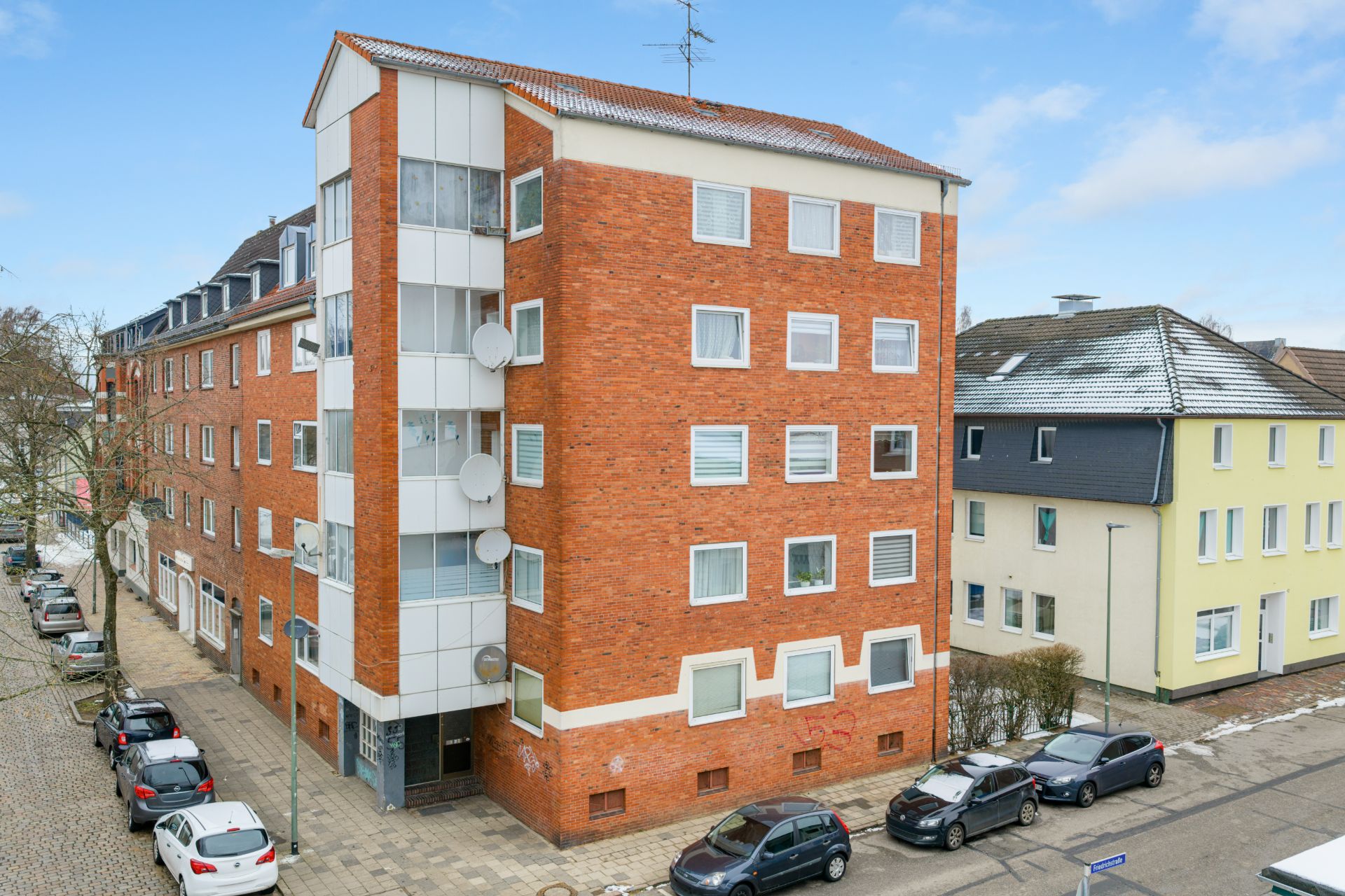 Vierstöckiges Wohnhaus aus rotem Backstein mit weißen Fenstern und parkenden Autos davor.