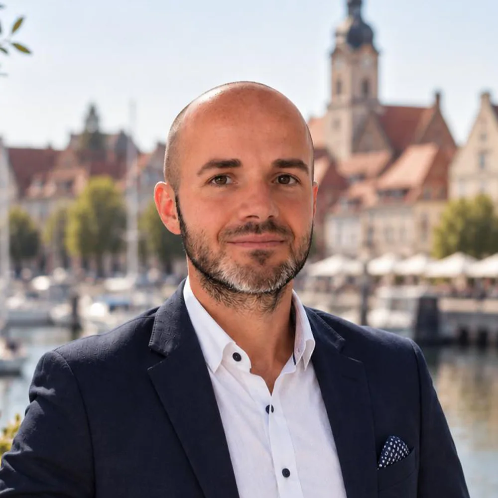 Ein Mann im Anzug steht draußen vor Wasser und historischen Gebäuden.