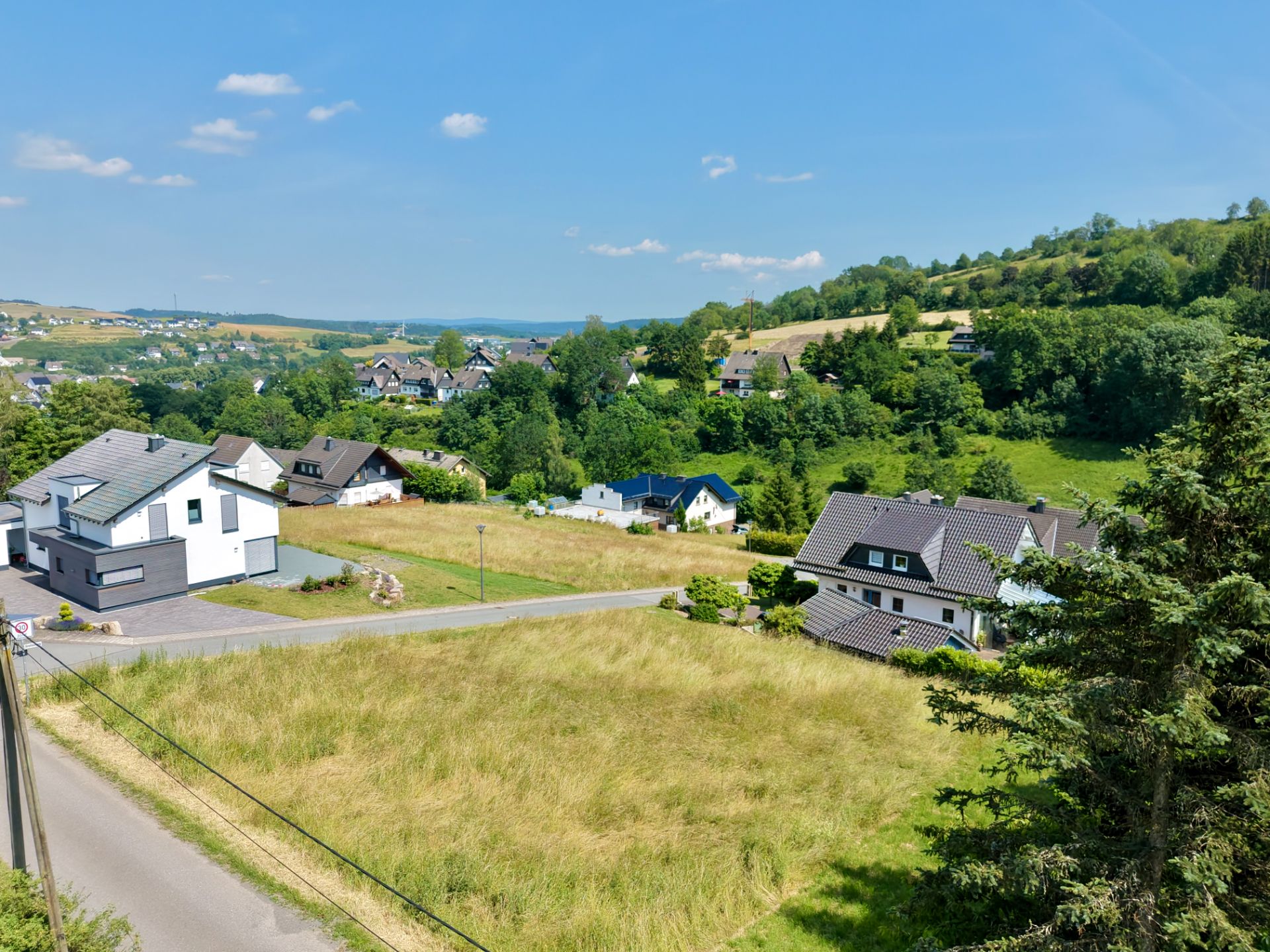Mehrere Einfamilienhäuser an einer ruhigen Straße mit Wiesen und Hügeln im Hintergrund.