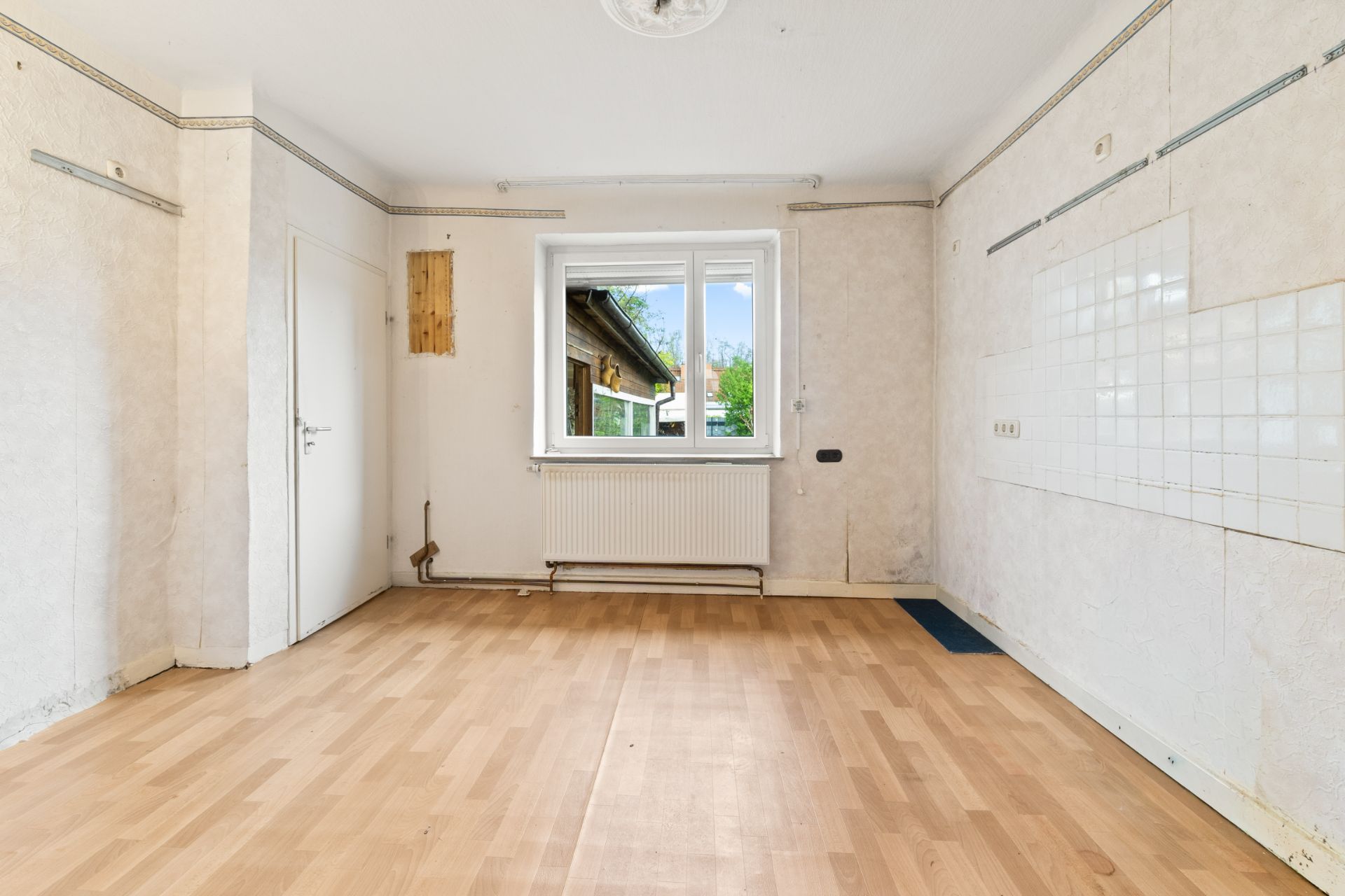 Leerer Raum mit hellem Laminatboden, Fenster, Heizung und weißen Wänden mit Fliesenspiegel.