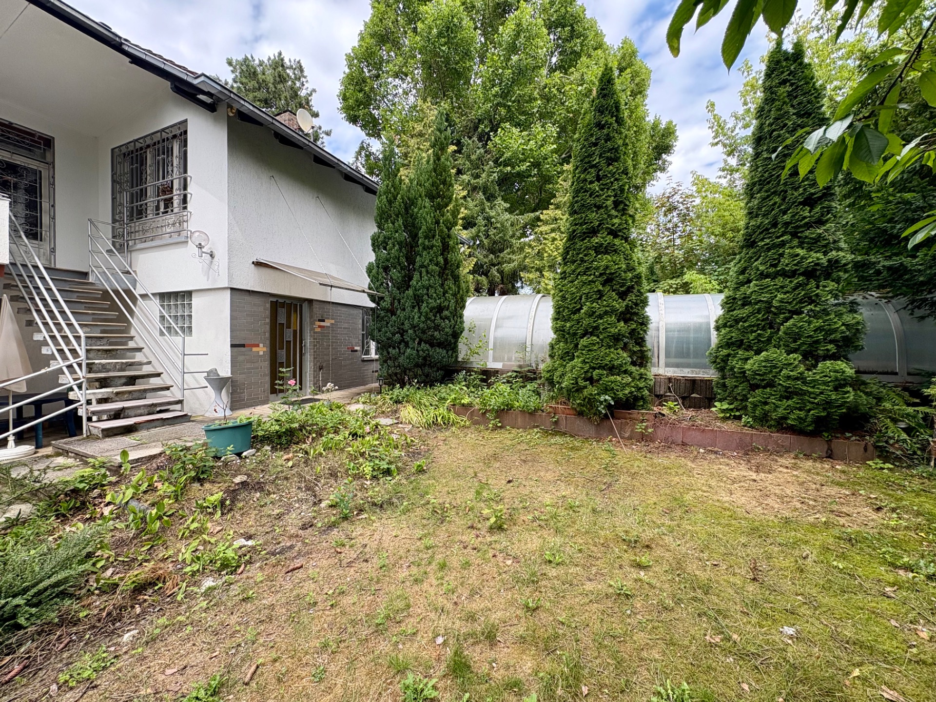 Garten mit Rasen, mehreren Bäumen und einem weißen Wohnhaus mit Außentreppe.