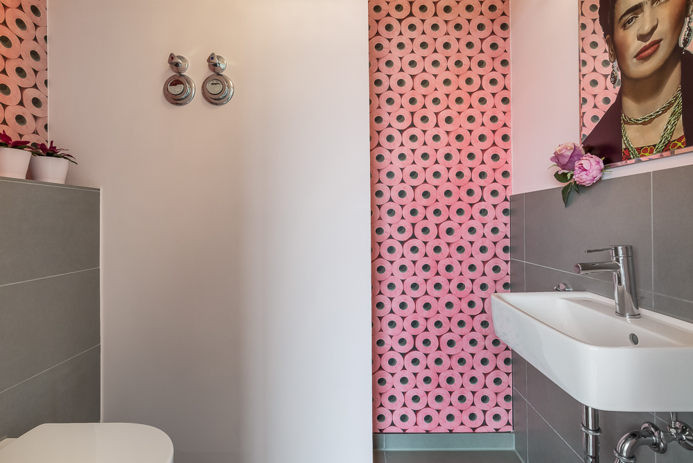 Kleines Bad mit grauen Fliesen, Waschbecken, Toilette und pink gemusterter Tapete.