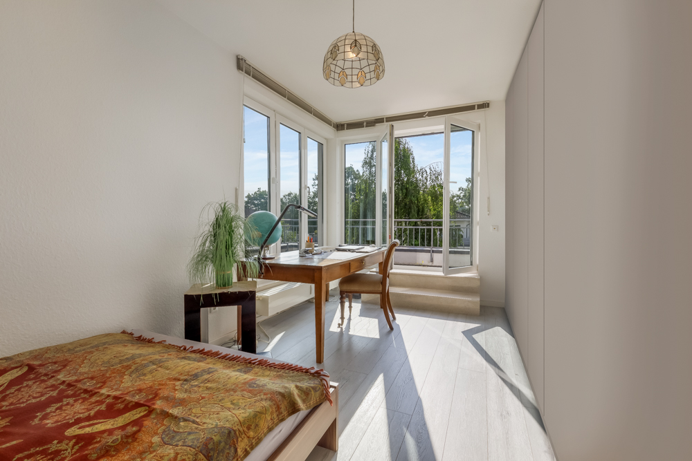 Helles Zimmer mit Bett, Schreibtisch, Pflanze und großen Fenstern sowie offenem Zugang zum Balkon.