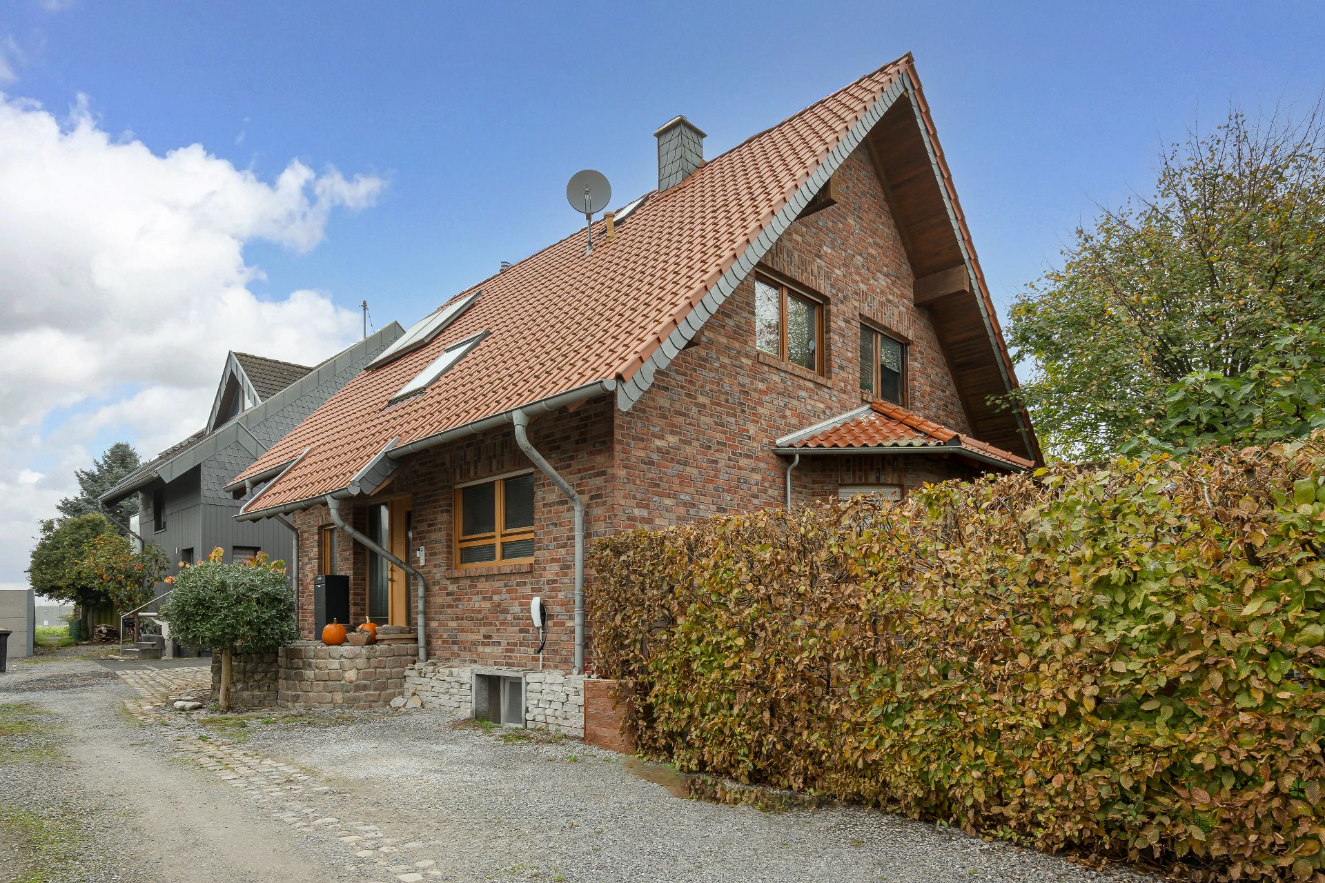 Backstein-Einfamilienhaus mit rotem Satteldach, Einfahrt und Hecke im Vordergrund.