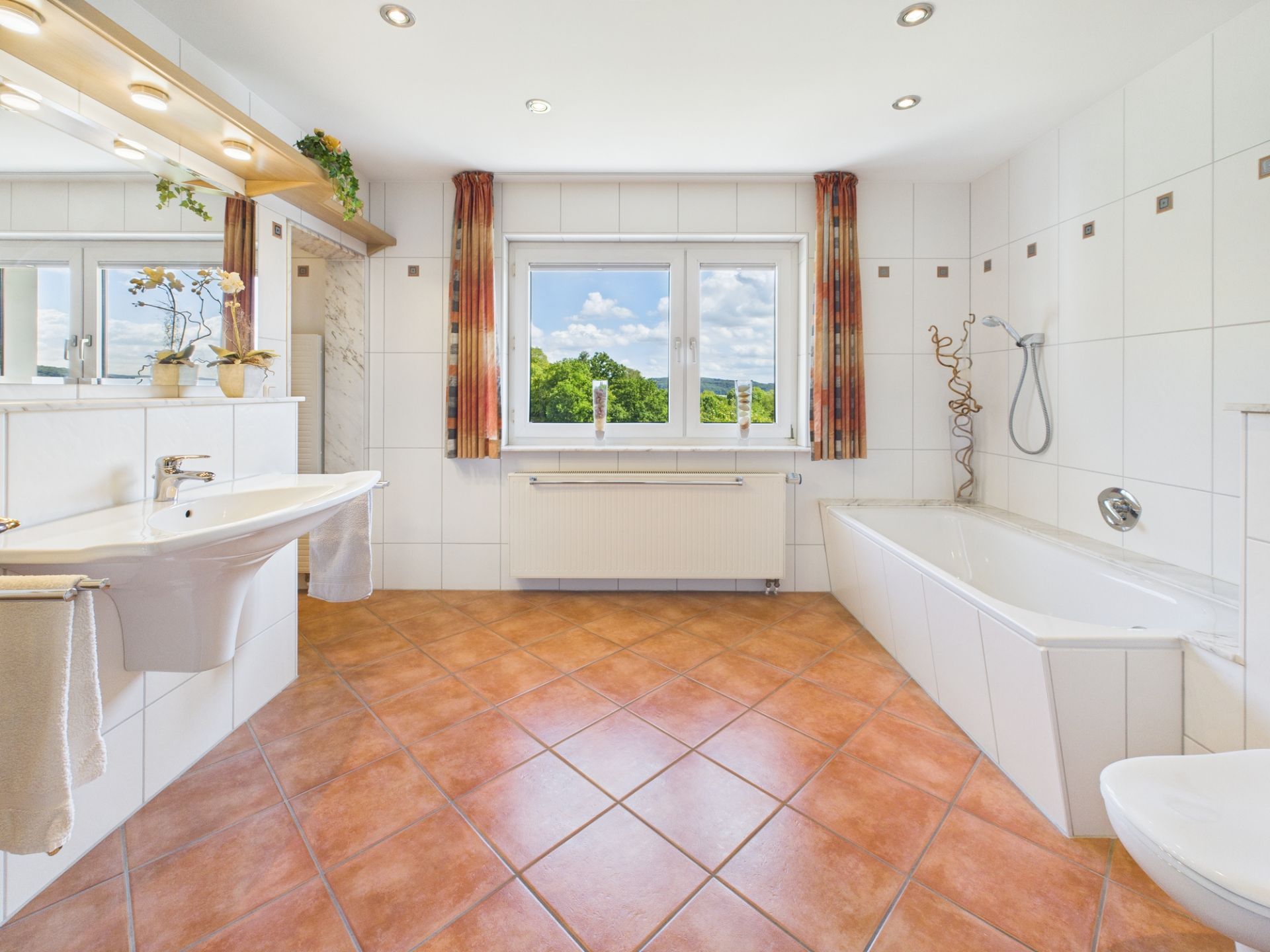 Helles Badezimmer mit Badewanne, Waschbecken, Spiegel und Fenster mit Blick ins Grüne.
