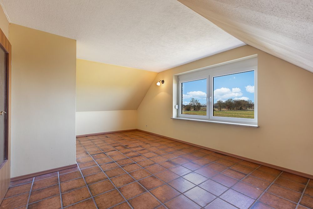 Leerer Raum mit schräger Decke, braunen Fliesen und großem Fenster mit Blick ins Grüne.
