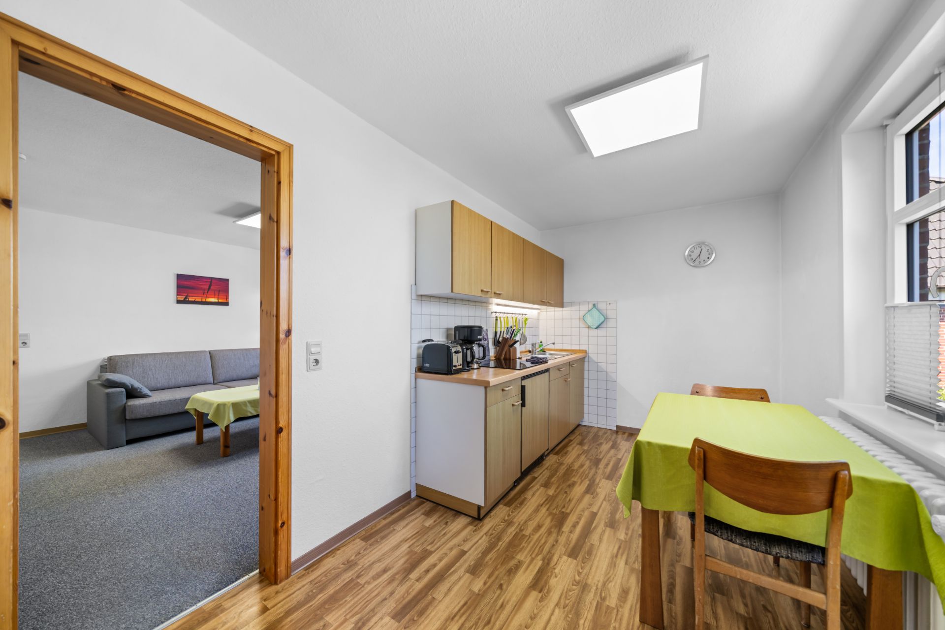 Küche mit Tisch und zwei Stühlen, Holzboden, Fenster und Zugang zu Wohnzimmer mit Sofa.