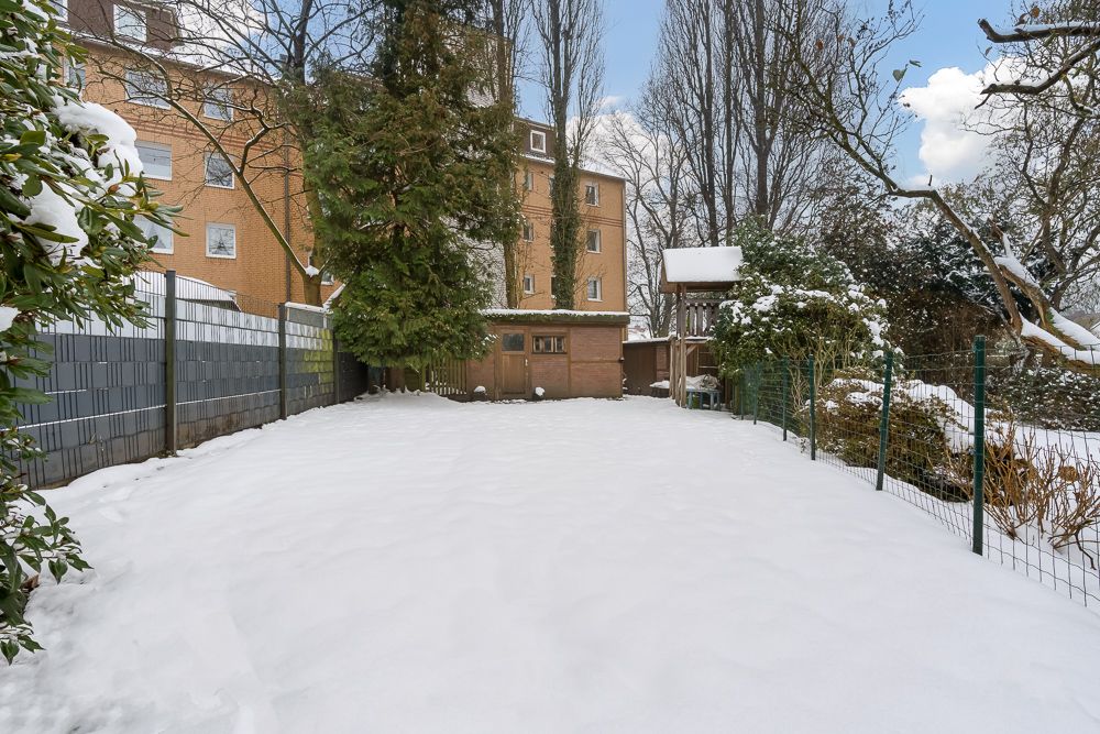 Schneebedeckter Garten mit Zaun, Sträuchern und Bäumen, hinten ein Mehrfamilienhaus.