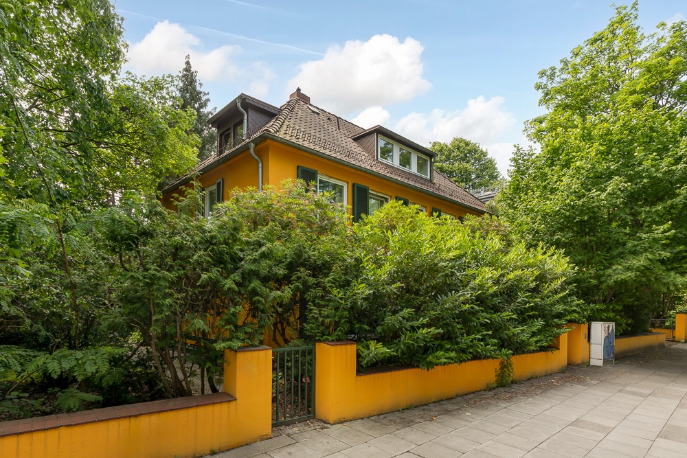 Gelbes Wohnhaus mit grünem Zaun und vielen Bäumen, von Gehweg aus sichtbar.