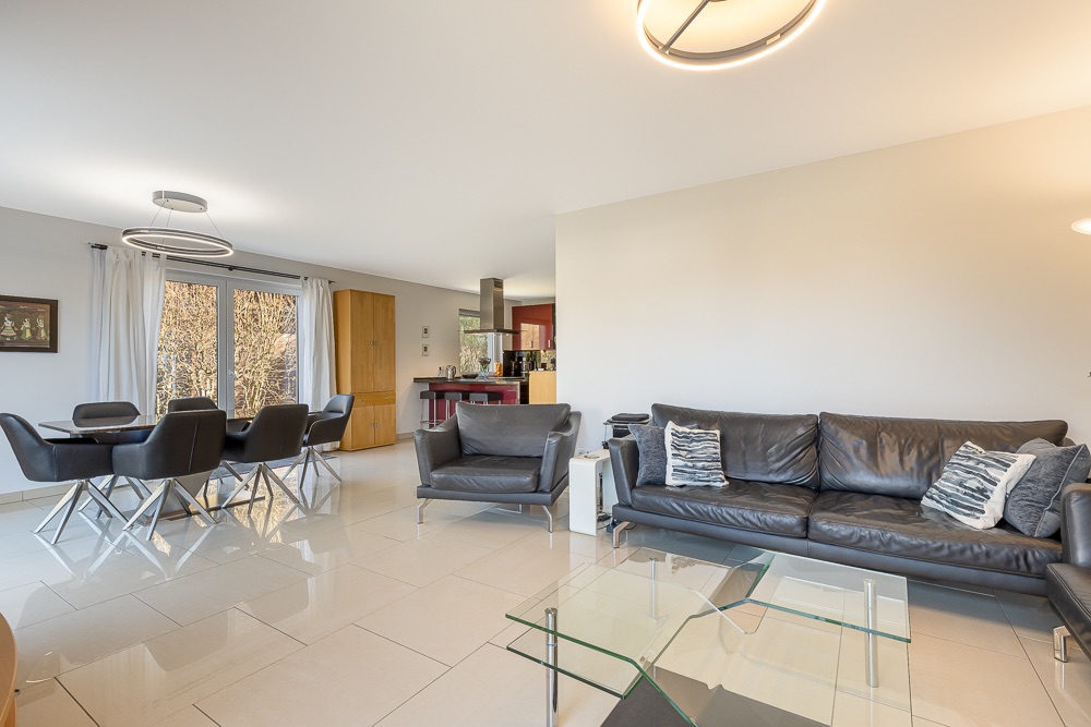 Helles Wohnzimmer mit schwarzem Sofa, Sessel, Glastisch und Essbereich mit sechs Stühlen.