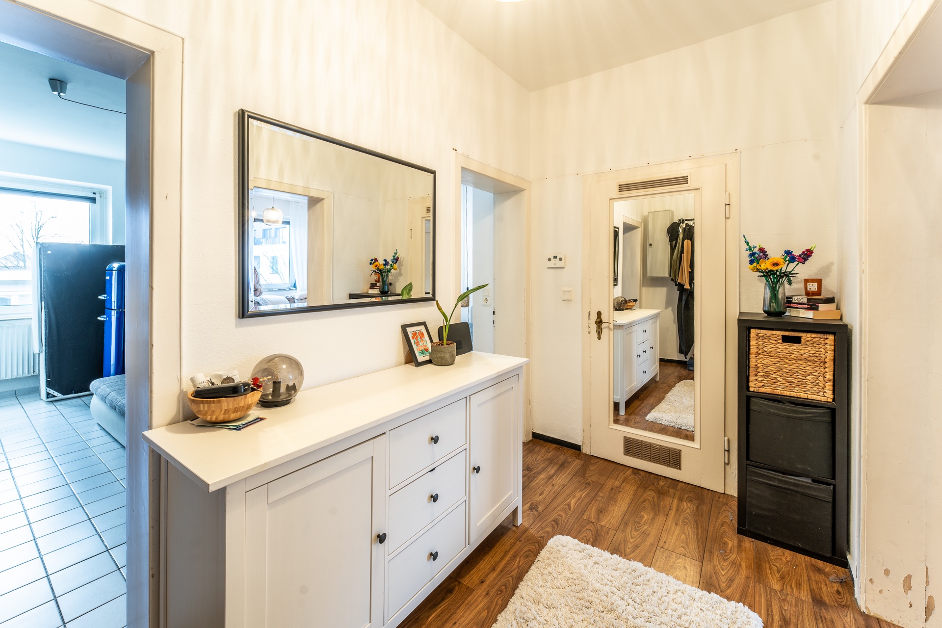 Heller Flur mit weißem Sideboard, großem Spiegel und Holzboden, daneben schwarze Kommode mit Blumen.