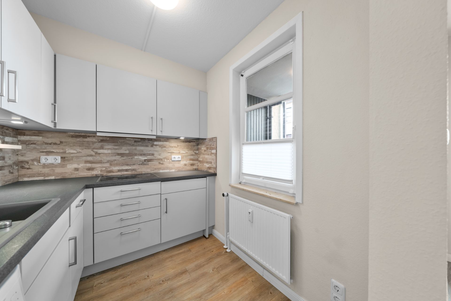 Moderne Küche mit weißen Schränken, Holzfußboden und Fenster mit Heizkörper darunter.