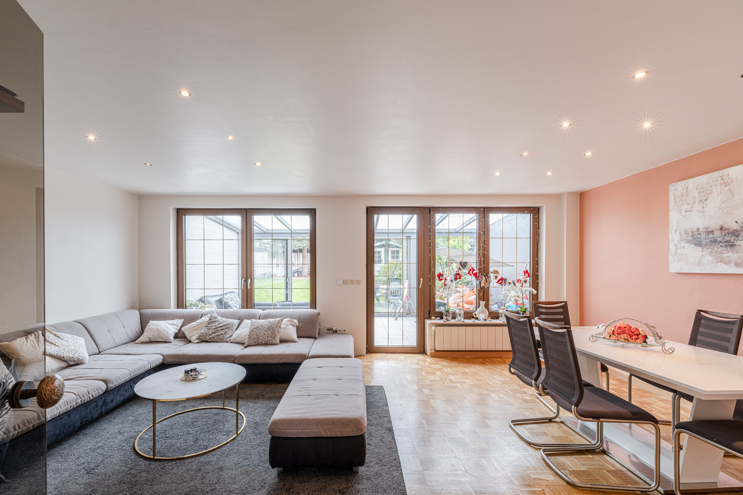 Helles Wohnzimmer mit Ecksofa, Couchtisch, Esstisch und großen Fenstern zum Garten.