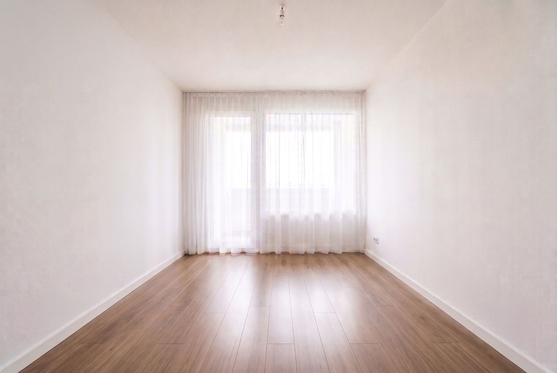Leerer, heller Raum mit Holzfußboden und großen Fenstern mit weißen Vorhängen.