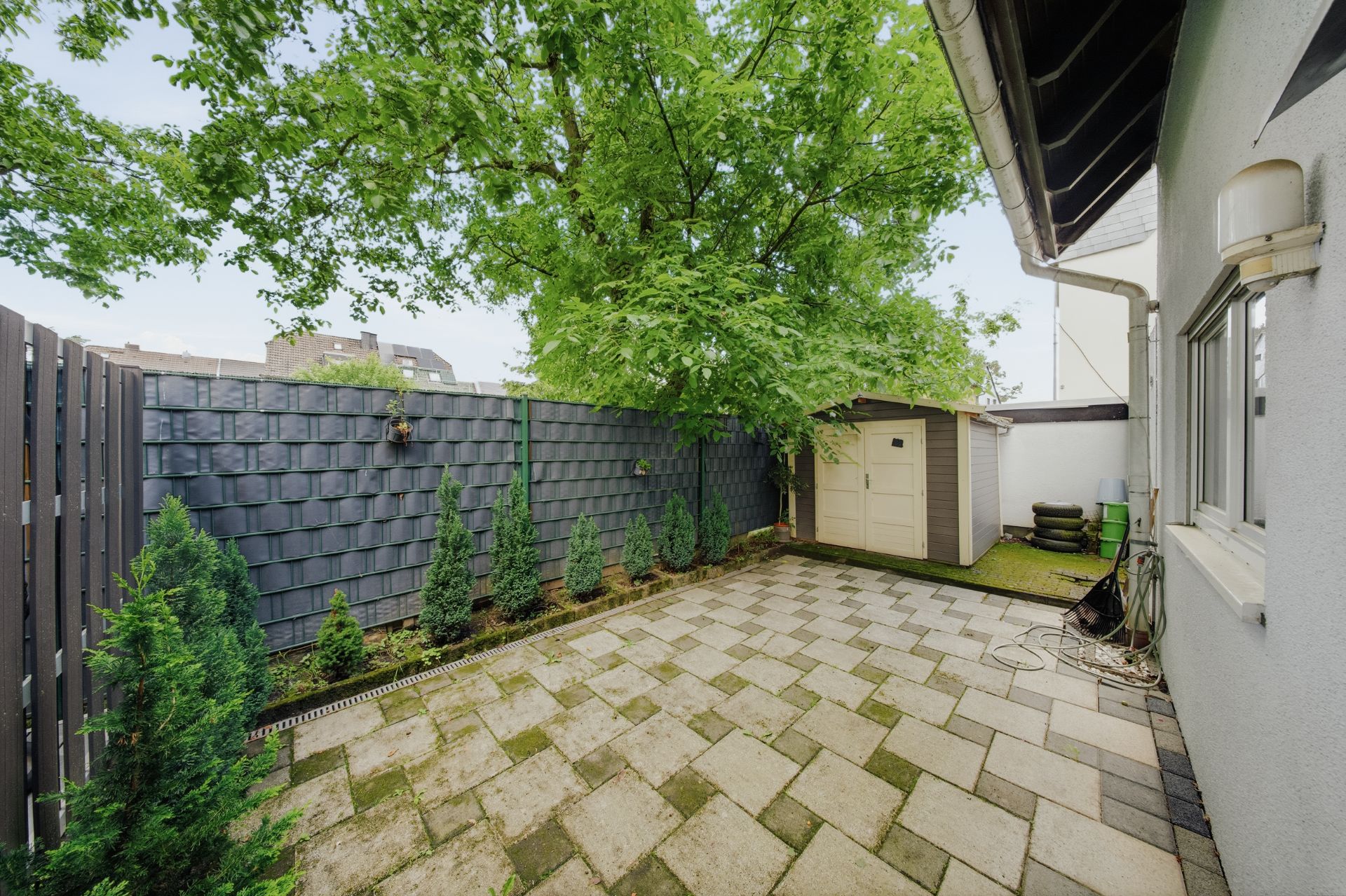 Kleiner, gepflasterter Innenhof mit Gartenhäuschen, Bäumen und angrenzender Hauswand.