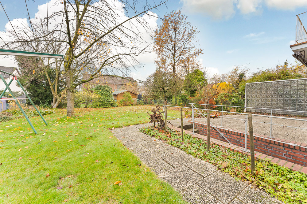 Garten mit Wiese, Bäumen, Gehweg und Metallgeländer neben einer kleinen Terrasse.