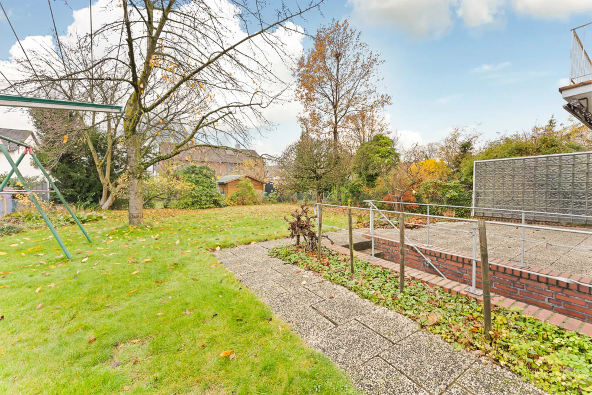 Garten mit Wiese, Bäumen, Gehweg und Metallgeländer neben einer kleinen Terrasse.