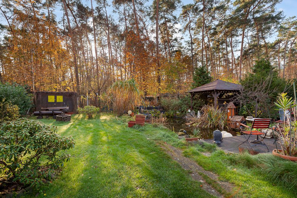 Garten mit Rasenfläche, Pavillon, Teich und Sitzbereich, umgeben von Bäumen.