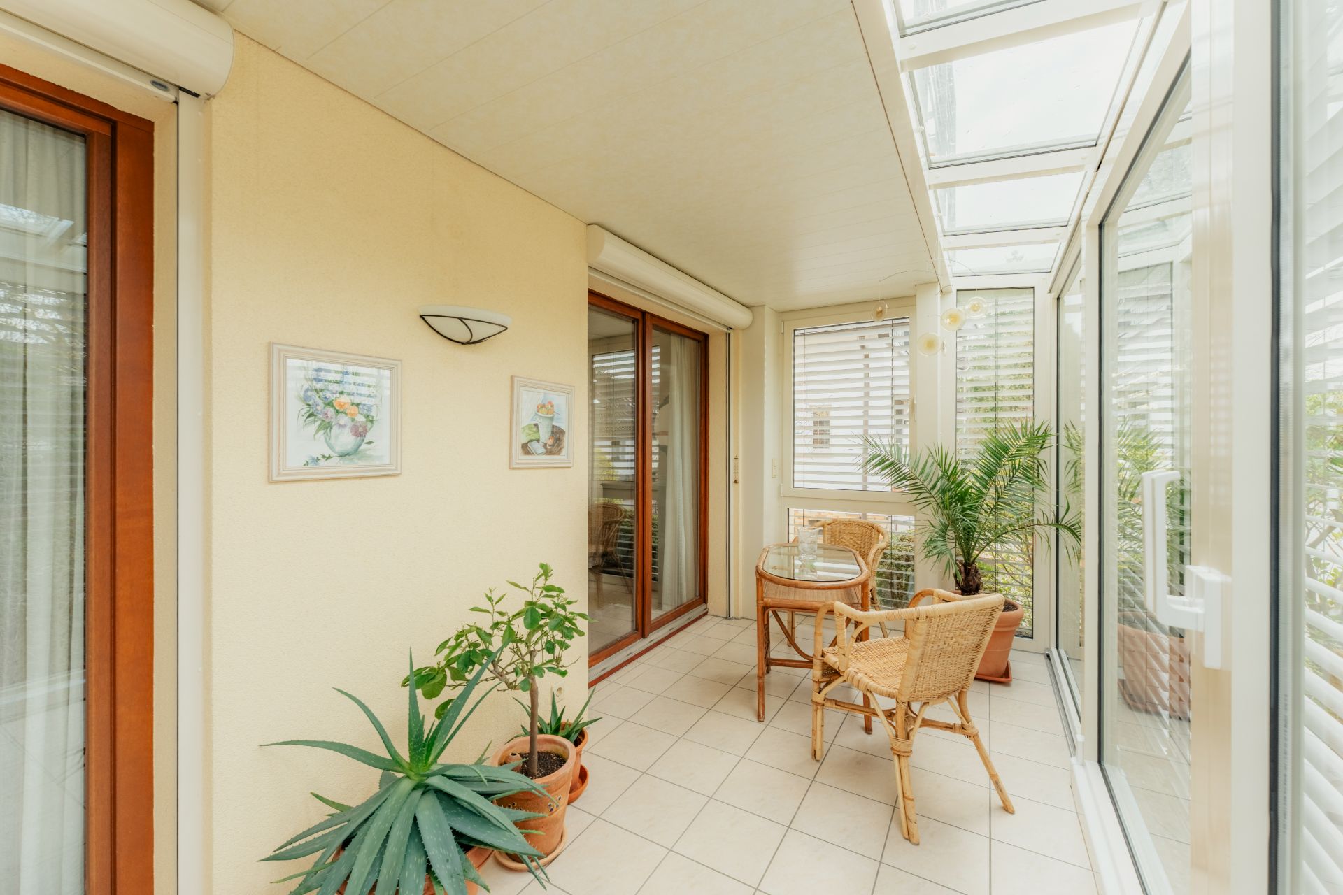 Heller Wintergarten mit Glasdach, Tisch, Stühlen, Topfpflanzen und Wandbildern.