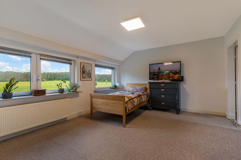 Helles Schlafzimmer mit Holzbett, Kommode, Fernseher und großen Fenstern mit Blick ins Grüne.