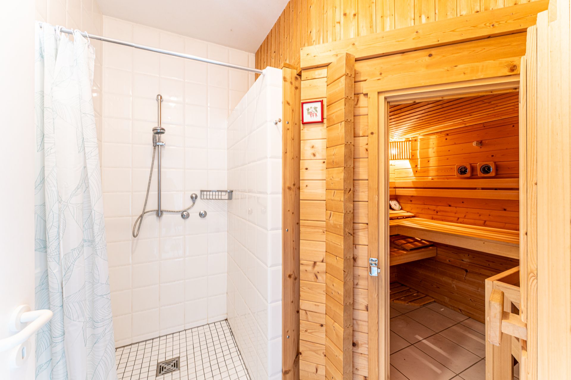 Duschbereich mit weißen Fliesen neben einer Holztür zu einer Sauna mit Holzbänken.