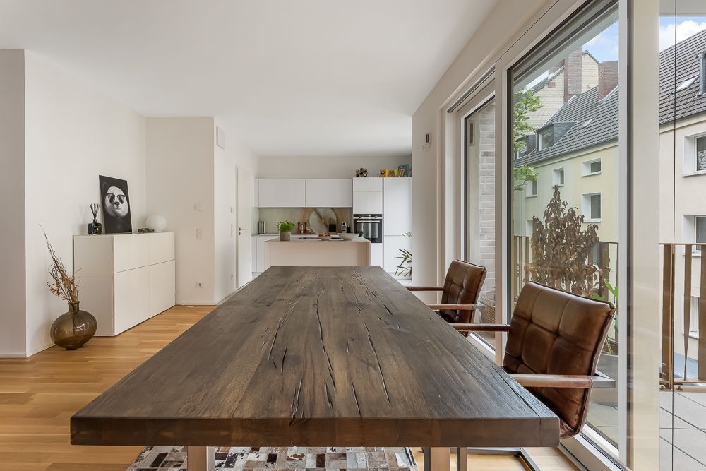Essbereich mit Holztisch und Lederstühlen vor großer Fensterfront, dahinter moderne Küche.