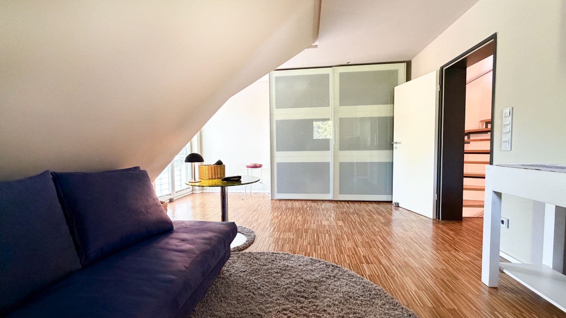 Helles Zimmer mit lila Sofa, rundem Teppich, Tisch und großem Schrank unter Dachschräge.