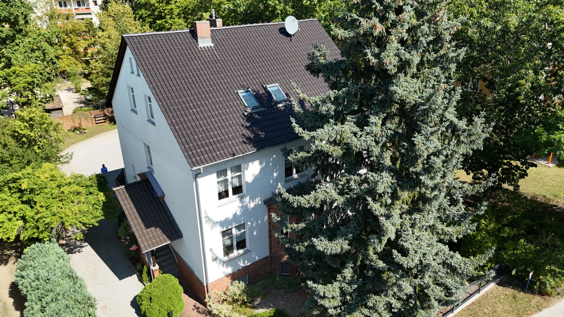 Zweistöckiges Wohnhaus mit dunklem Dach, vielen Fenstern und großen Bäumen im Garten.