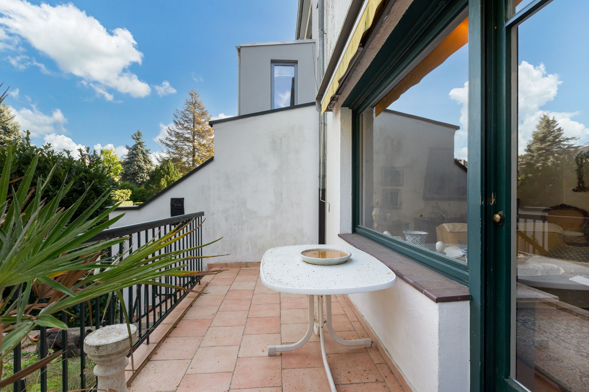 Balkon mit Fliesenboden, einem weißen Tisch und Blick auf Garten und Bäume.