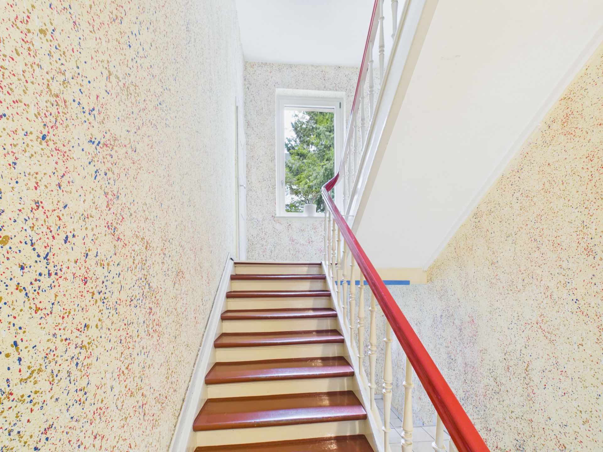 Treppenhaus mit bunten Wänden und rotem Handlauf, Fenster mit Blick auf Bäume.