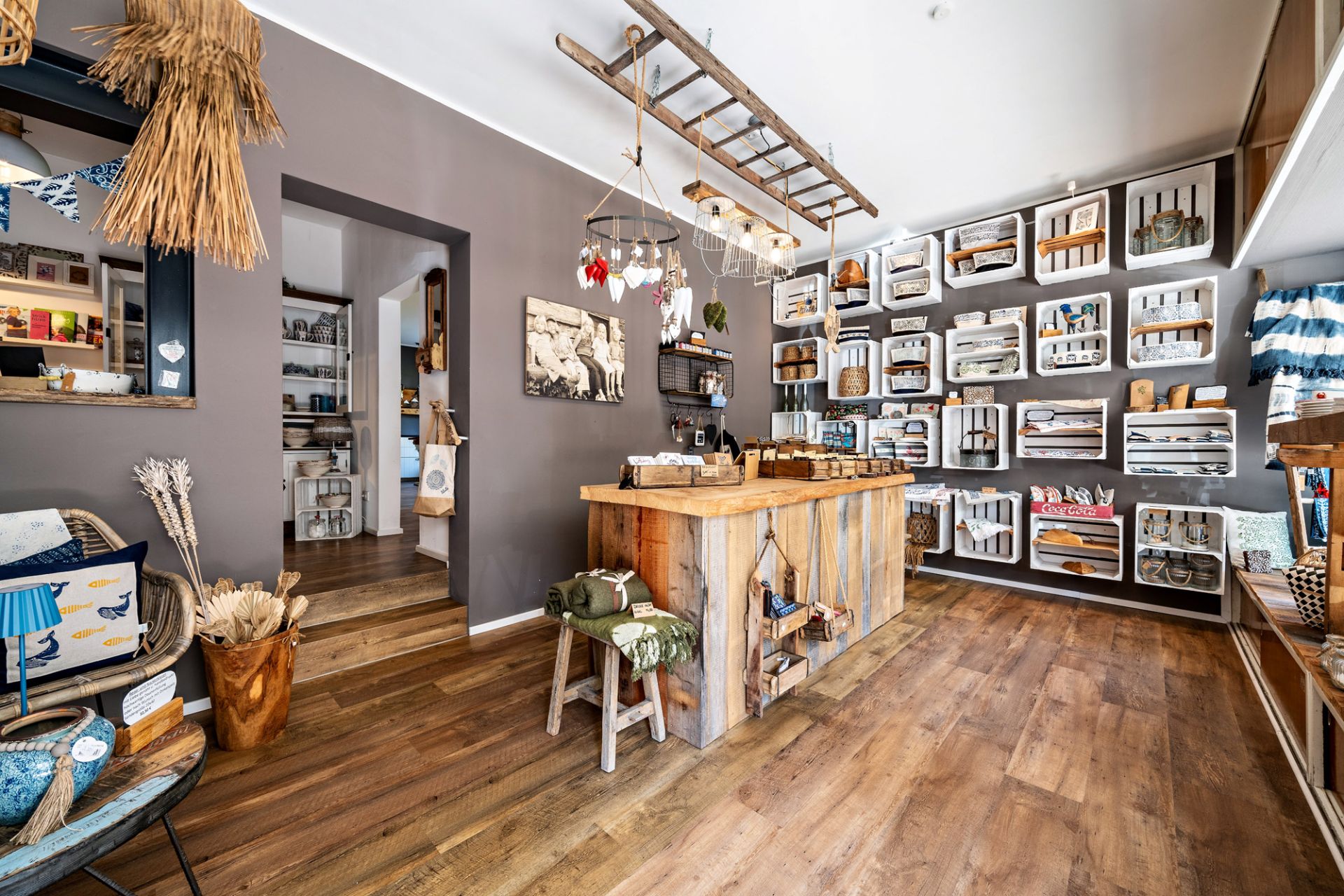 Heller Raum mit Holzboden, großem Holztisch und Regalen mit Dekoartikeln an der Wand.