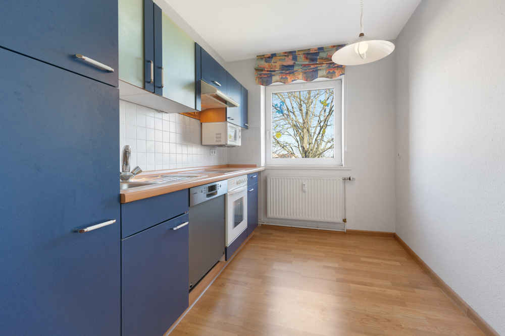 Küche mit blauen Schränken, Herd, Spüle und Fenster mit Blick auf einen Baum.